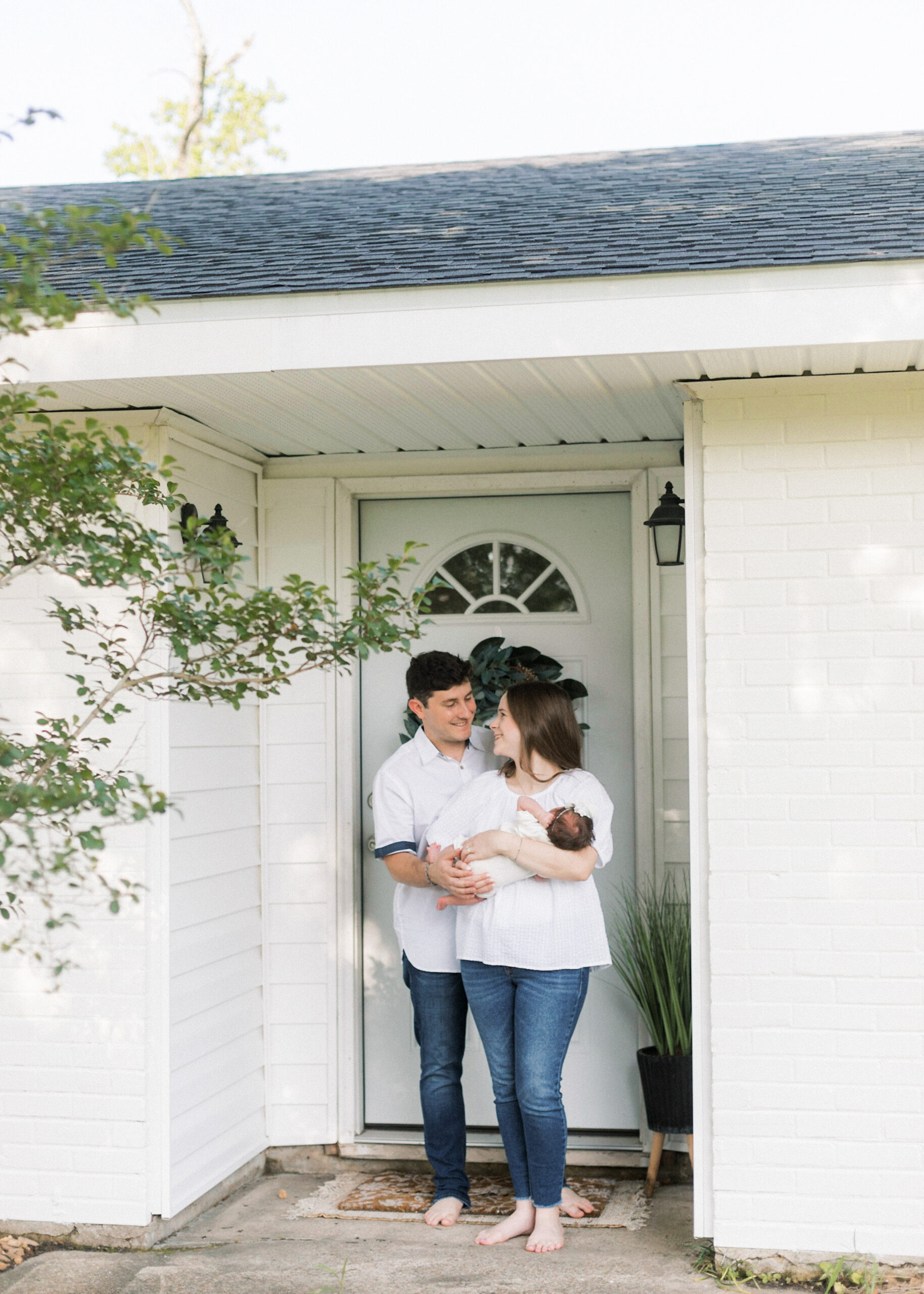 Adorable parents embrace their precious bundle of joy, standing proudly in front of their home's entrance during a heartwarming in-home newborn photoshoot